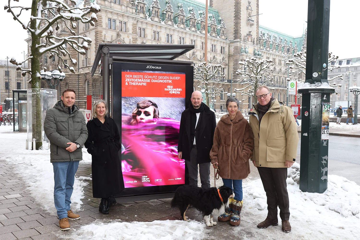 Fünf Personen und ein Hund stehen rund um ein Digital City Light Poster an einer Bus-Wartehalle am Hamburger Rathaus. Das Digital City Light Poster zeigt eine Kampagne, die auf den plötzlichen Tod durch Epilepsie aufmerksam macht.
