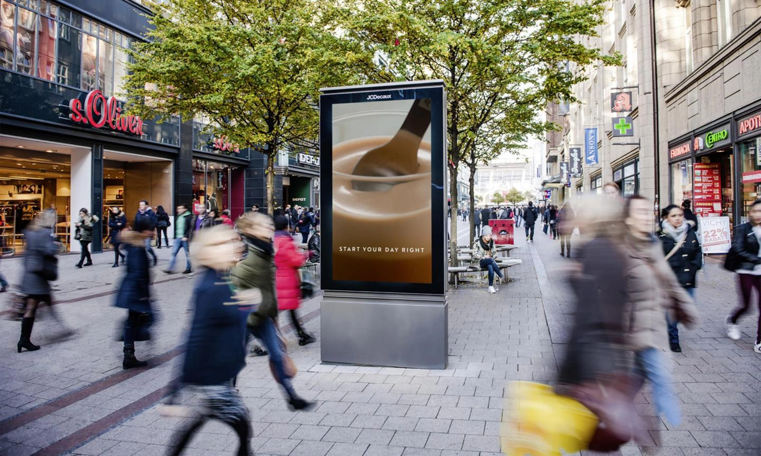 Digital City Light Poster mit Kaffee-Werbung in einer belebten Einkaufsstraße in Hamburg, umgeben von Geschäften und vorbeigehenden Menschen.