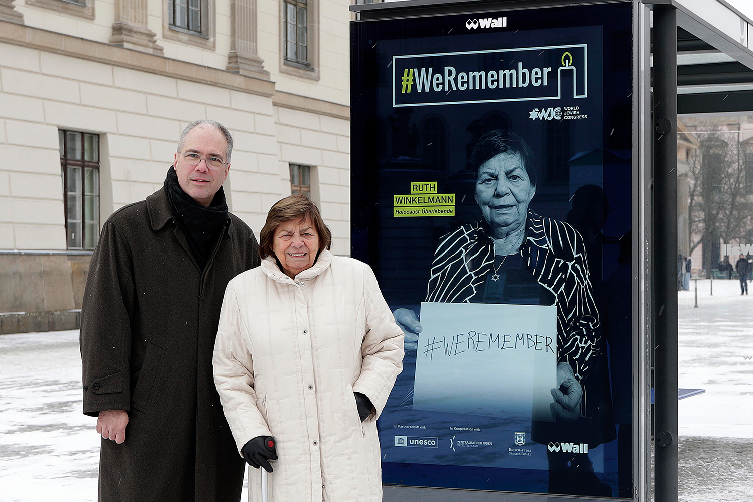 Portrait von Patrick Möller und Ruth Winkelmann vor einem Digital City Light Poster, dass Ruth Winkelmann als ein Gesicht der WeRemember-Kampagne zeigt.