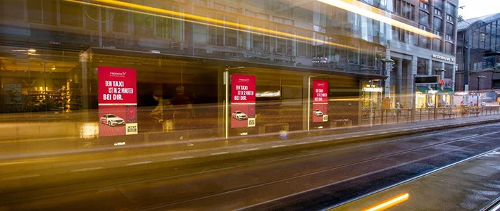 Drei Digital City Light Poster zeigen Fahrdienst-Werbung in Berliner Friedrichstraße bei Dämmerung.