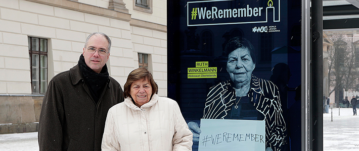 Portrait von Patrick Möller und Ruth Winkelmann vor einem Digital City Light Poster, dass Ruth Winkelmann als ein Gesicht der WeRemember-Kampagne zeigt.